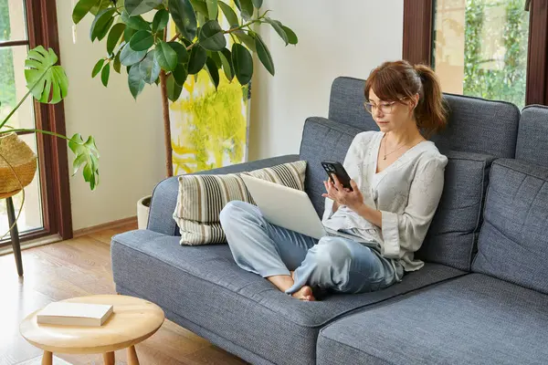 Bir kadın evinde rahat bir şekilde kanepede çalışır, bilgisayarını kullanır ve telefonda konuşur, bitkilerle ve doğal ışıkla çevrili..