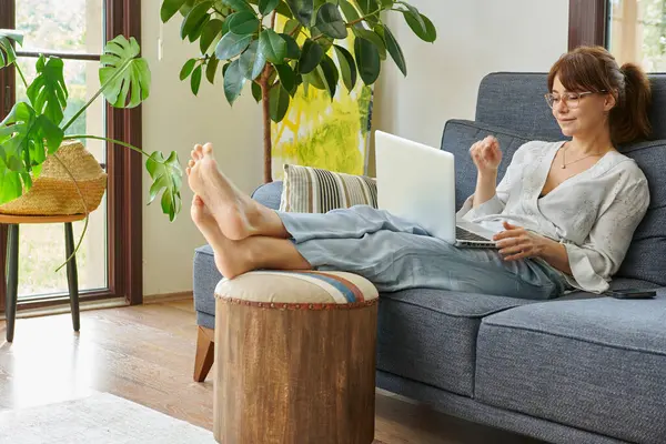 Bir kadın evinde rahat bir şekilde kanepede çalışır, bilgisayarını kullanır ve telefonda konuşur, bitkilerle ve doğal ışıkla çevrili..