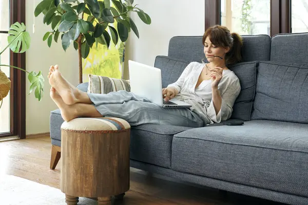 Bir kadın evinde rahat bir şekilde kanepede çalışır, bilgisayarını kullanır ve telefonda konuşur, bitkilerle ve doğal ışıkla çevrili..