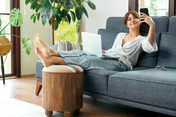 Bir kadın evinde rahat bir şekilde kanepede çalışır, bilgisayarını kullanır ve telefonda konuşur, bitkilerle ve doğal ışıkla çevrili..