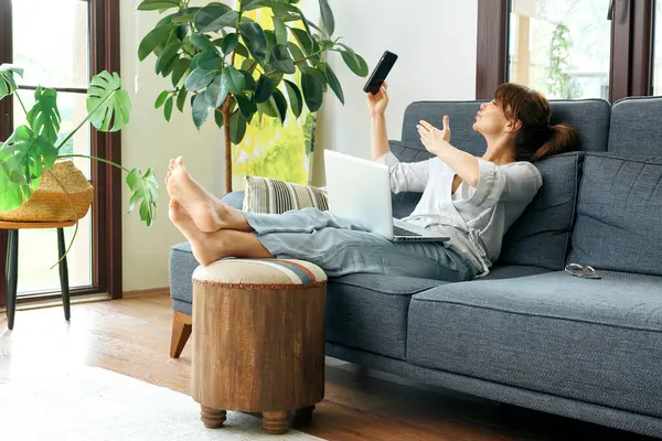 Bir kadın evinde rahat bir şekilde kanepede çalışır, bilgisayarını kullanır ve telefonda konuşur, bitkilerle ve doğal ışıkla çevrili..