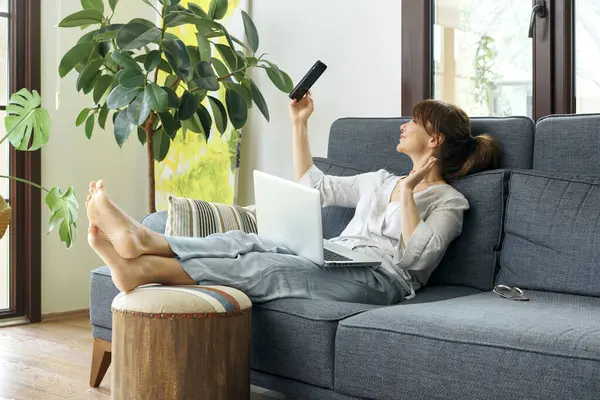 Bir kadın evinde rahat bir şekilde kanepede çalışır, bilgisayarını kullanır ve telefonda konuşur, bitkilerle ve doğal ışıkla çevrili..