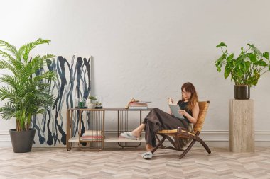 Bir kadın rahat rahat kitap okuyor şık bir rattan şezlongda, yanında kitap ve dekor tutan bir sehpa ile..