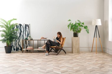 Bir kadın rahat rahat kitap okuyor şık bir rattan şezlongda, yanında kitap ve dekor tutan bir sehpa ile..