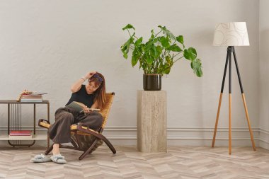 Bir kadın rahat rahat kitap okuyor şık bir rattan şezlongda, yanında kitap ve dekor tutan bir sehpa ile..