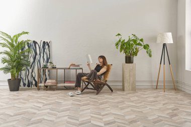 Bir kadın rahat rahat kitap okuyor şık bir rattan şezlongda, yanında kitap ve dekor tutan bir sehpa ile..