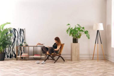 Bir kadın rahat rahat kitap okuyor şık bir rattan şezlongda, yanında kitap ve dekor tutan bir sehpa ile..