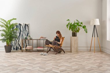 Bir kadın rahat rahat kitap okuyor şık bir rattan şezlongda, yanında kitap ve dekor tutan bir sehpa ile..