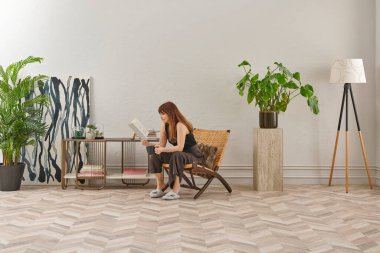 Bir kadın rahat rahat kitap okuyor şık bir rattan şezlongda, yanında kitap ve dekor tutan bir sehpa ile..