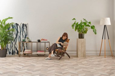 Bir kadın rahat rahat kitap okuyor şık bir rattan şezlongda, yanında kitap ve dekor tutan bir sehpa ile..