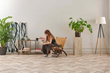 Görüntü, örülmüş sandalyeye rahatça oturmuş tablete bakan bir kadını gösteriyor. Rahat bir ev ortamı izlenimi uyandırmak için evde rahat terlikler giyiyor..