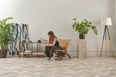 Görüntü, örülmüş sandalyeye rahatça oturmuş tablete bakan bir kadını gösteriyor. Rahat bir ev ortamı izlenimi uyandırmak için evde rahat terlikler giyiyor..