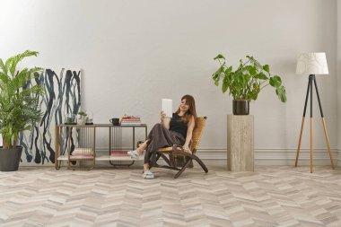 Görüntü, örülmüş sandalyeye rahatça oturmuş tablete bakan bir kadını gösteriyor. Rahat bir ev ortamı izlenimi uyandırmak için evde rahat terlikler giyiyor..