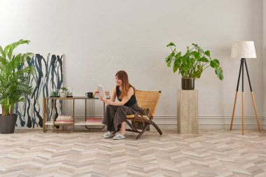 Görüntü, örülmüş sandalyeye rahatça oturmuş tablete bakan bir kadını gösteriyor. Rahat bir ev ortamı izlenimi uyandırmak için evde rahat terlikler giyiyor..