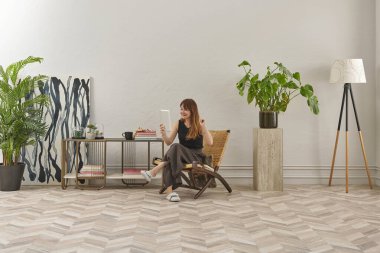 Görüntü, örülmüş sandalyeye rahatça oturmuş tablete bakan bir kadını gösteriyor. Rahat bir ev ortamı izlenimi uyandırmak için evde rahat terlikler giyiyor..