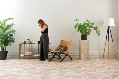 Görüntü, örülmüş sandalyeye rahatça oturmuş tablete bakan bir kadını gösteriyor. Rahat bir ev ortamı izlenimi uyandırmak için evde rahat terlikler giyiyor..