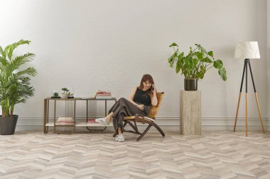 Görüntü, örülmüş sandalyeye rahatça oturmuş tablete bakan bir kadını gösteriyor. Rahat bir ev ortamı izlenimi uyandırmak için evde rahat terlikler giyiyor..