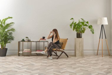 Görüntü, örülmüş sandalyeye rahatça oturmuş tablete bakan bir kadını gösteriyor. Rahat bir ev ortamı izlenimi uyandırmak için evde rahat terlikler giyiyor..