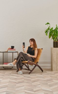 Görüntü, örülmüş sandalyeye rahatça oturmuş tablete bakan bir kadını gösteriyor. Rahat bir ev ortamı izlenimi uyandırmak için evde rahat terlikler giyiyor..