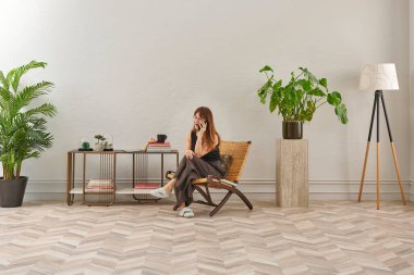 Görüntü, örülmüş sandalyeye rahatça oturmuş tablete bakan bir kadını gösteriyor. Rahat bir ev ortamı izlenimi uyandırmak için evde rahat terlikler giyiyor..