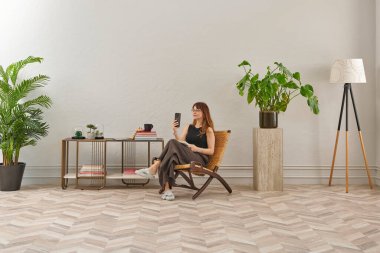 Görüntü, örülmüş sandalyeye rahatça oturmuş tablete bakan bir kadını gösteriyor. Rahat bir ev ortamı izlenimi uyandırmak için evde rahat terlikler giyiyor..