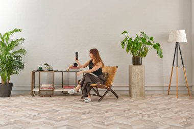 Görüntü, örülmüş sandalyeye rahatça oturmuş tablete bakan bir kadını gösteriyor. Rahat bir ev ortamı izlenimi uyandırmak için evde rahat terlikler giyiyor..