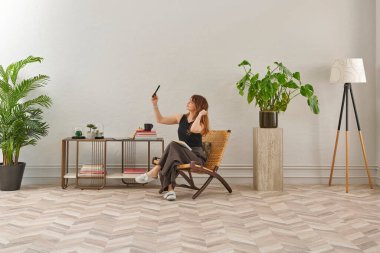 Görüntü, örülmüş sandalyeye rahatça oturmuş tablete bakan bir kadını gösteriyor. Rahat bir ev ortamı izlenimi uyandırmak için evde rahat terlikler giyiyor..