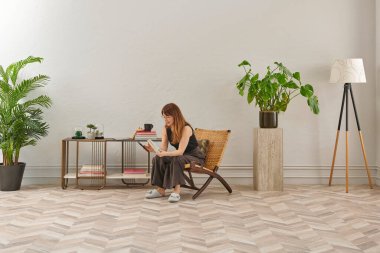 Gözlüklü bir kadın dokuma sandalyede oturur ve telefona bakar. Siyah bir bluz, siyah pantolon ve rahat gri terlikler giyiyor..