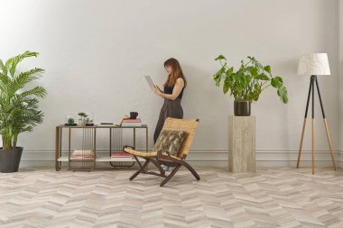 Gözlüklü bir kadın dokuma sandalyede oturur ve telefona bakar. Siyah bir bluz, siyah pantolon ve rahat gri terlikler giyiyor..