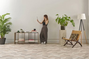 Gözlüklü bir kadın dokuma sandalyede oturur ve telefona bakar. Siyah bir bluz, siyah pantolon ve rahat gri terlikler giyiyor..