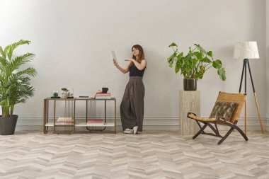 Gözlüklü bir kadın dokuma sandalyede oturur ve telefona bakar. Siyah bir bluz, siyah pantolon ve rahat gri terlikler giyiyor..