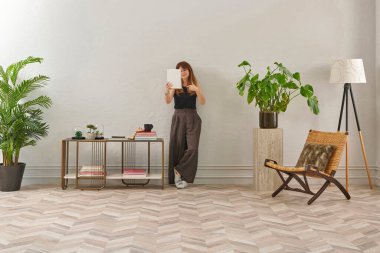 Gözlüklü bir kadın dokuma sandalyede oturur ve telefona bakar. Siyah bir bluz, siyah pantolon ve rahat gri terlikler giyiyor..