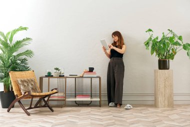 Gözlüklü bir kadın dokuma sandalyede oturur ve telefona bakar. Siyah bir bluz, siyah pantolon ve rahat gri terlikler giyiyor..