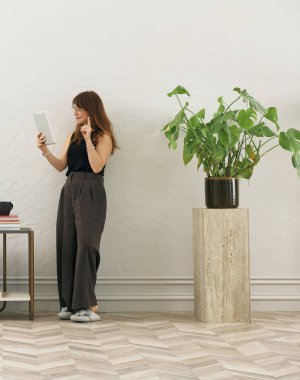 Gözlüklü bir kadın dokuma sandalyede oturur ve telefona bakar. Siyah bir bluz, siyah pantolon ve rahat gri terlikler giyiyor..