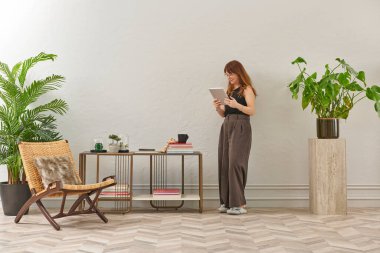 Gözlüklü bir kadın dokuma sandalyede oturur ve telefona bakar. Siyah bir bluz, siyah pantolon ve rahat gri terlikler giyiyor..