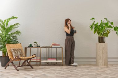 Gözlüklü bir kadın dokuma sandalyede oturur ve telefona bakar. Siyah bir bluz, siyah pantolon ve rahat gri terlikler giyiyor..