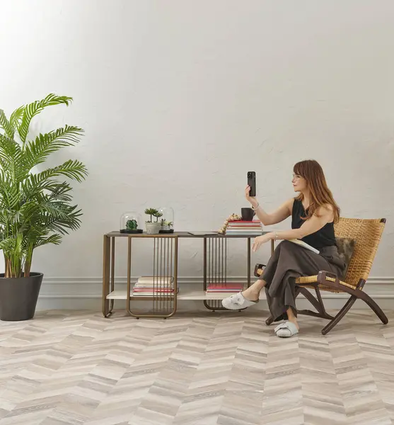 Görüntü, örülmüş sandalyeye rahatça oturmuş tablete bakan bir kadını gösteriyor. Rahat bir ev ortamı izlenimi uyandırmak için evde rahat terlikler giyiyor..