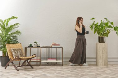 Gözlüklü bir kadın dokuma sandalyede oturur ve telefona bakar. Siyah bir bluz, siyah pantolon ve rahat gri terlikler giyiyor..