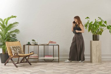 Gözlüklü bir kadın dokuma sandalyede oturur ve telefona bakar. Siyah bir bluz, siyah pantolon ve rahat gri terlikler giyiyor..