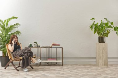 Gözlüklü bir kadın dokuma sandalyede oturur ve telefona bakar. Siyah bir bluz, siyah pantolon ve rahat gri terlikler giyiyor..