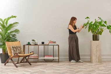 Siyah bluzlu ve koyu renkli geniş pantolonlu bir kadın büyük bir bitkiyi sular. Bitki koyu tencerede açık renkli bir kaidede oturur..