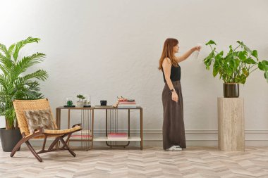 Siyah bluzlu ve koyu renkli geniş pantolonlu bir kadın büyük bir bitkiyi sular. Bitki koyu tencerede açık renkli bir kaidede oturur..