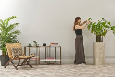 Siyah bluzlu ve koyu renkli geniş pantolonlu bir kadın büyük bir bitkiyi sular. Bitki koyu tencerede açık renkli bir kaidede oturur..