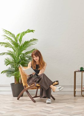 Bir kadın evde kitap okuyor, telefon ve tablete bakıyor. Bitkinin dekoratif iç stili ve vazosu.