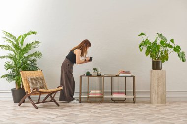 Siyah bluzlu ve koyu renkli geniş pantolonlu bir kadın büyük bir bitkiyi sular. Bitki koyu tencerede açık renkli bir kaidede oturur..