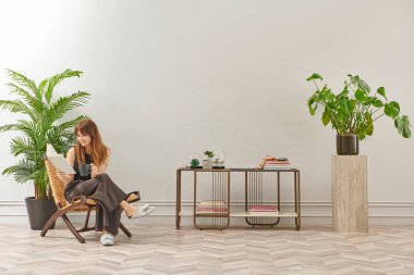 Bir kadın evde kitap okuyor, telefon ve tablete bakıyor. Bitkinin dekoratif iç stili ve vazosu.