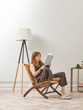 Bir kadın evde kitap okuyor, telefon ve tablete bakıyor. Bitkinin dekoratif iç stili ve vazosu.