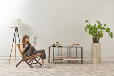 Bir kadın evde kitap okuyor, telefon ve tablete bakıyor. Bitkinin dekoratif iç stili ve vazosu.