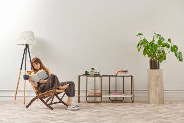 Bir kadın evde kitap okuyor, telefon ve tablete bakıyor. Bitkinin dekoratif iç stili ve vazosu.