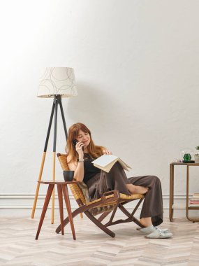 Bir kadın evde kitap okuyor, telefon ve tablete bakıyor. Bitkinin dekoratif iç stili ve vazosu.